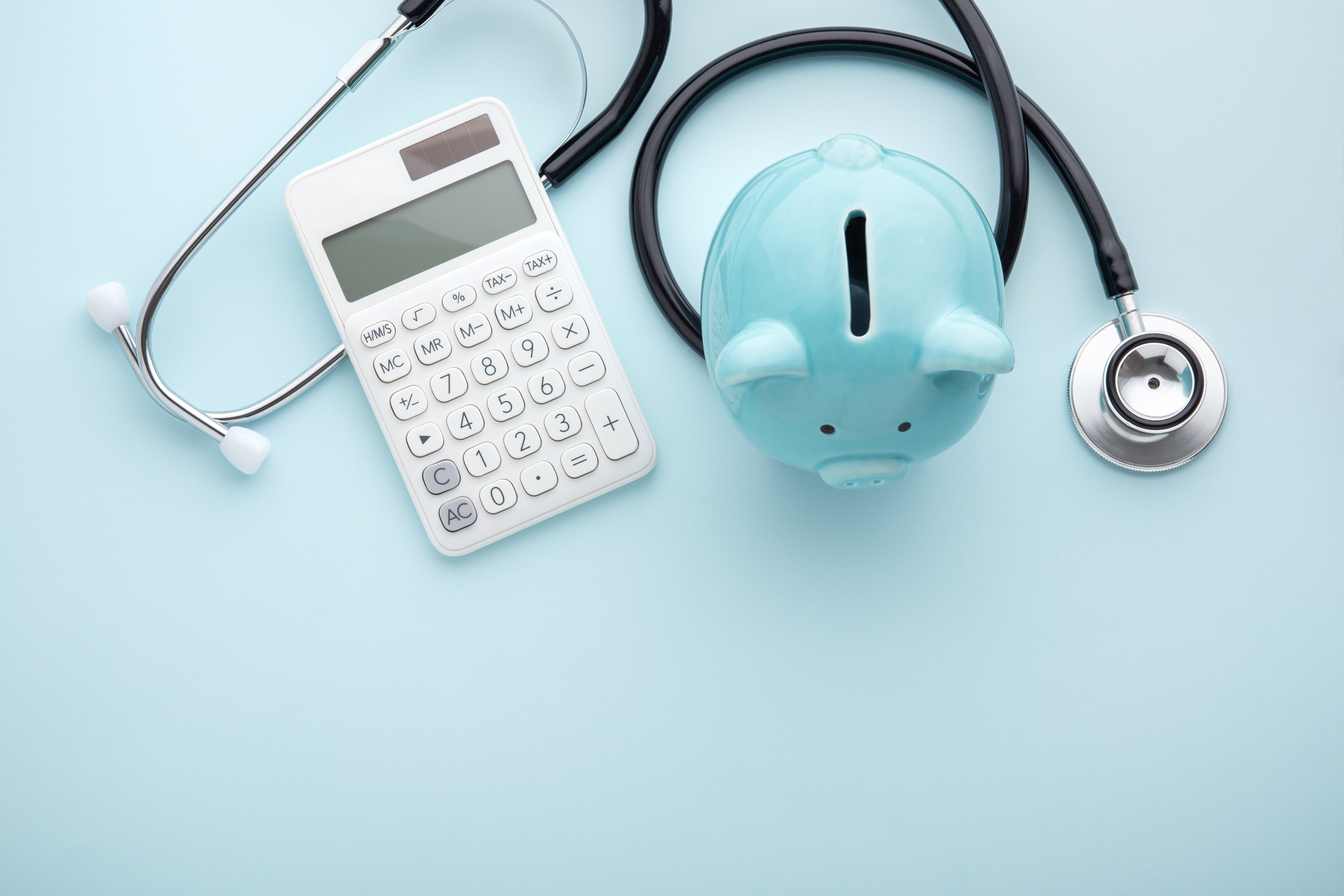 A calculator, a light blue piggy bank, and a stethoscope are arranged on a light blue surface, symbolizing financial planning and healthcare costs.