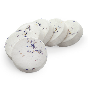 Four round white bath or body soaps with dried purple and yellow botanical flecks, arranged in a fan shape on a solid white background.