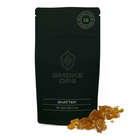 A black stand-up pouch labeled SMOKE OPS SHATTER with amber crystalline product pieces placed in front on a white background.