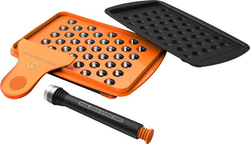 A set of accessories for filling dosing capsules, including a filling device, spatula, plunger, grinder, magazine with sealed capsules, and a scale. The grinder and magazine have orange accents.