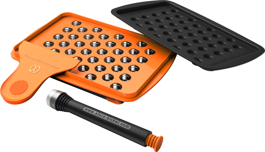 A set of accessories for filling dosing capsules, including a filling device, spatula, plunger, grinder, magazine with sealed capsules, and a scale. The grinder and magazine have orange accents.